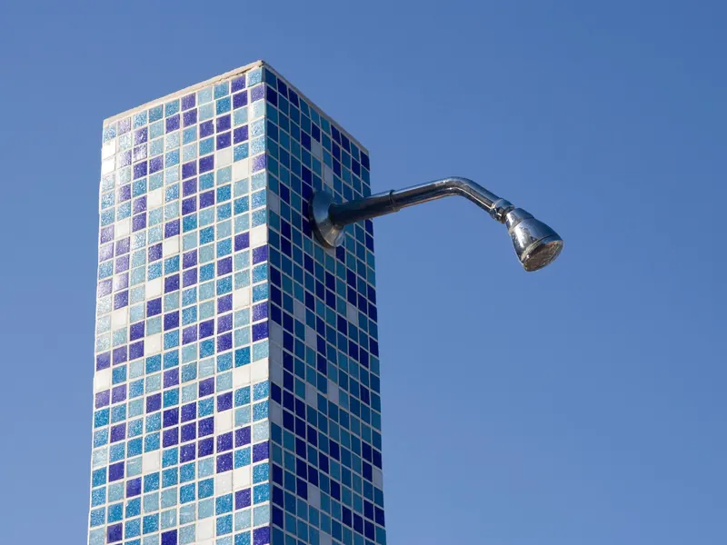 An outdoor shower head mounted on a tiled column against a clear sky