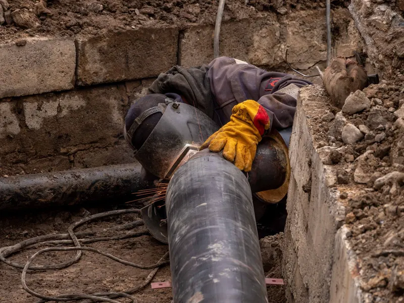 A worker in an open trench repairing a large underground pipe